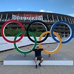 Grad Andrew Molina sits outdoors with his arms spread in front of a set of Olympic rings. There is a stadium with a banner for the 2020 Tokyo Olympics behind him.